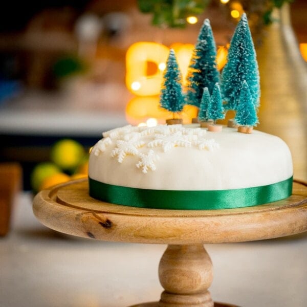 Square image showing a simple decorated Christmas cake - with a green ribbon surrounding the cake, white fondant snow flakes and mini Christmas trees on top. The cake is on a wooden cake stand on a kitchen surface with glowy lights and a bowl of fruit in the background.