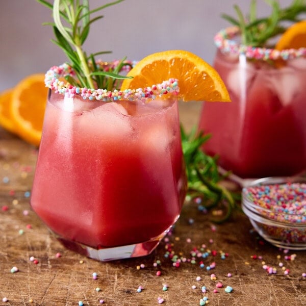 A square shot of two glasses of Christmas Margarita placed on a wooden surface. In the left of the background, there are a few slices of orange. In between the two glasses, there is a sprig of fresh rosemary, and in front of the glass on the right, there is a small glass dish of rainbow sprinkles. This is all set on a light grey background.