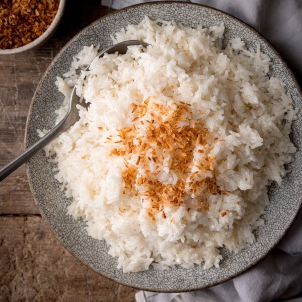 A square, overhead shot of Stovetop Coconut Rice. The rice is on a light grey plate in the centre of the image there is a silver spoon sticking out of the left side of the dish. To the right of the plate, there is a light grey napkin tucked beneath the plate. To the top left of the plate, you can see the corner of a white ceramic mug filled with toasted shredded coconut flakes. This is all set on a wooden surface.