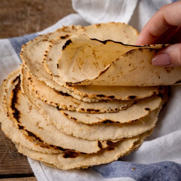 A square shot of corn tortillas. The image shows 9 tortillas stacked on top of each other. The top tortilla is being folded in half by a hand coming from the right-hand side of the screen. Beneath the stack of tortillas, there is a white and blue stripy napkin. This is all set on a wooden surface.