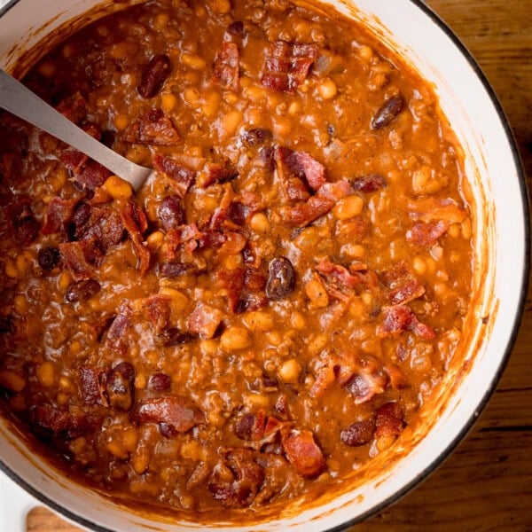 Square, close-up, overhead image of cowboy beans in a white casserole dish with a metal spoon sticking out.