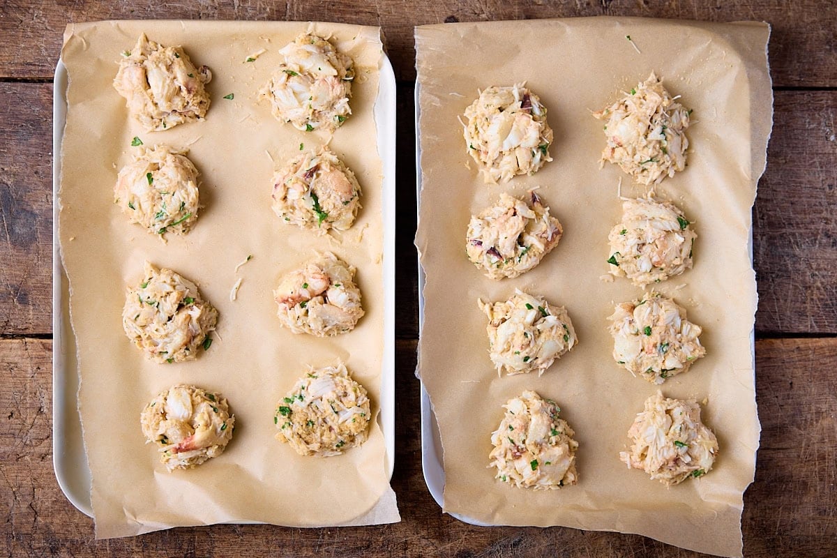 Overhead image of 16 uncooked mini crab cakes on baking parchment on top of two trays.