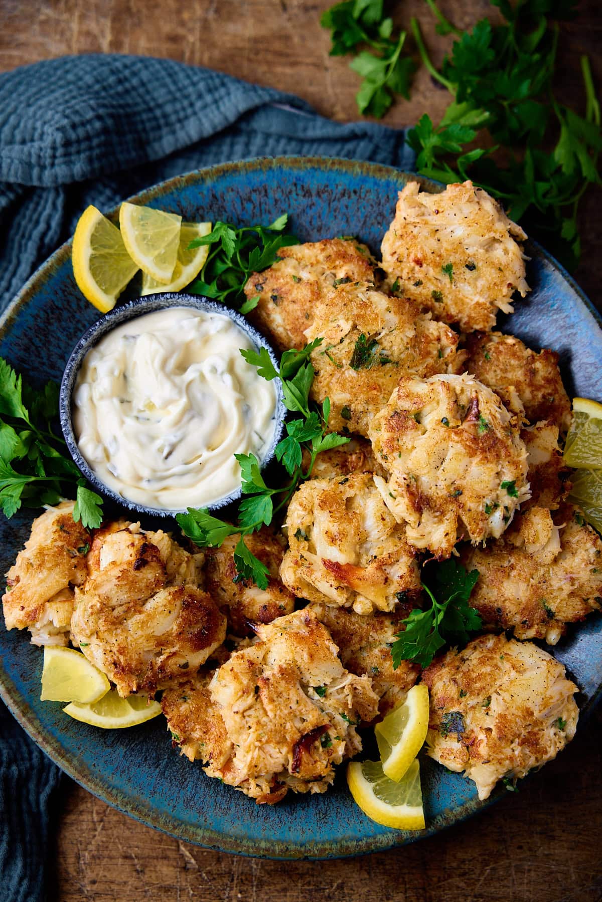 Overhead tall image of a plate of Maryland-style crab cakes on a mottled blue plate. There is a small bowl of tartar sauce, some lemon slices and fresh parsley also on the plate. The plate in on a wooden background and there is more parsley and a teal napkin near the plate.