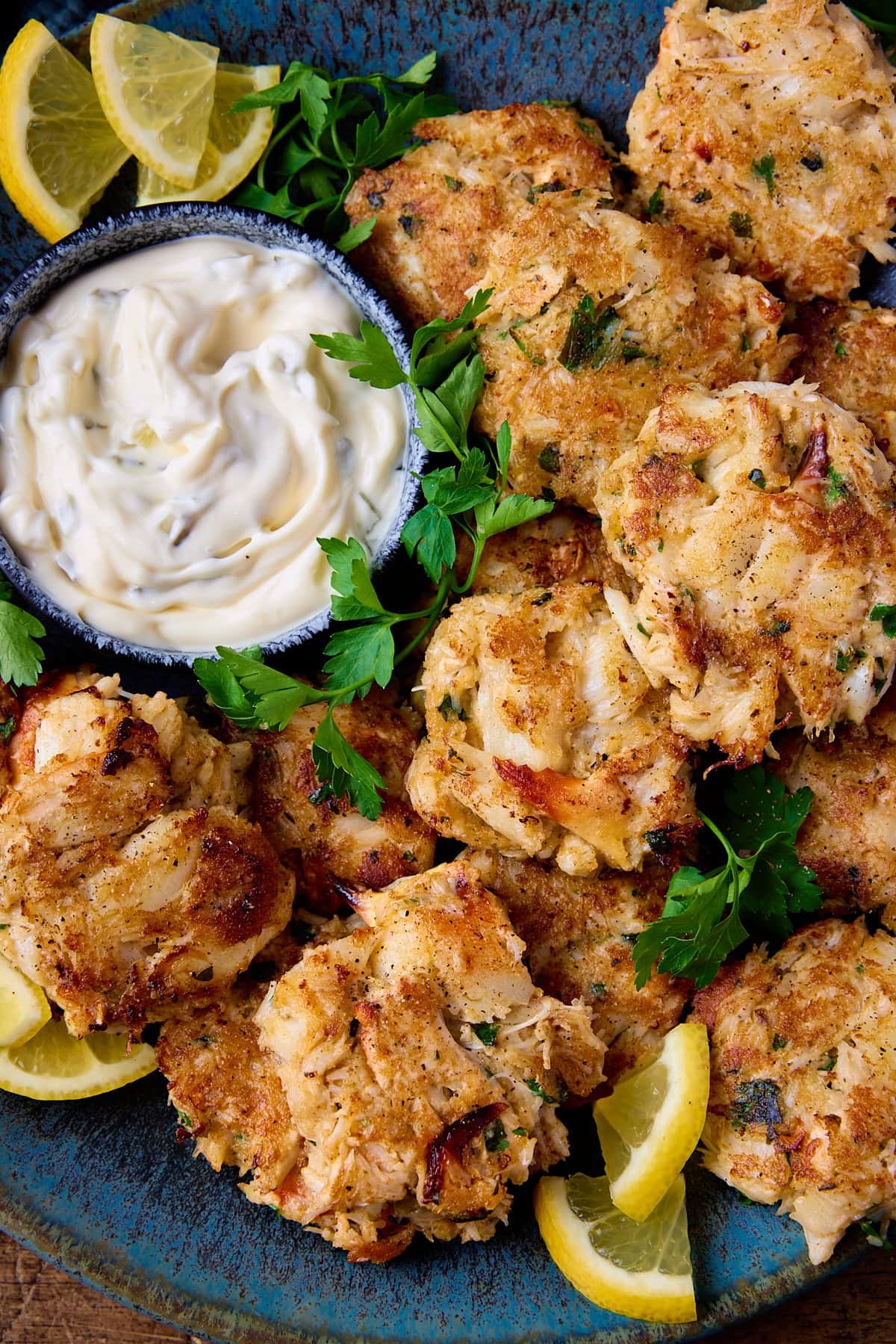 Overhead close-up image of a plate of Maryland-style crab cakes on a mottled blue plate. There is a small bowl of tartar sauce, some lemon slices and fresh parsley also on the plate.