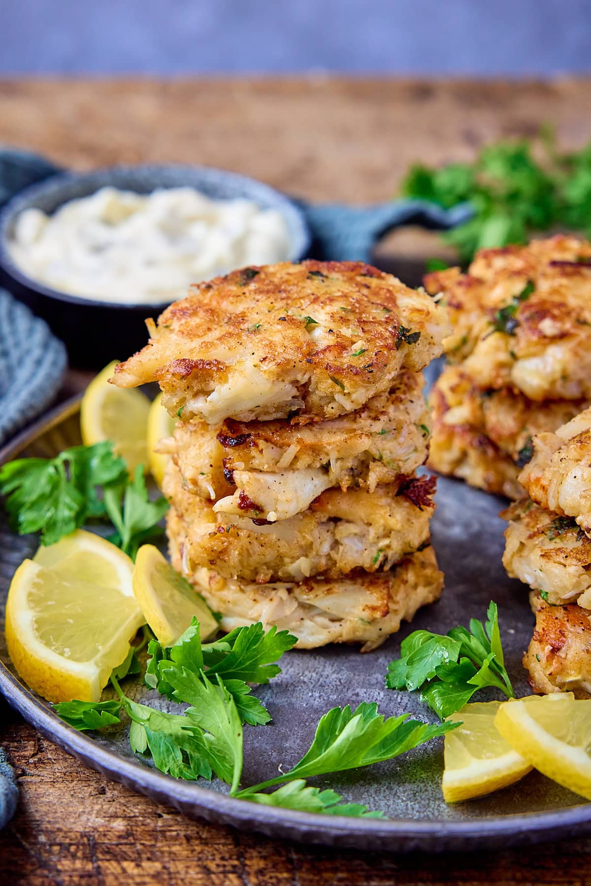 Tall side-on image of Maryland-style crab cakes in small piles on a grey plate. There are some lemon slices and fresh parsley also on the plate. The plate in on a wooden background and there is more parsley, a small bowl of tartar sauce and a light green napkin near the plate.