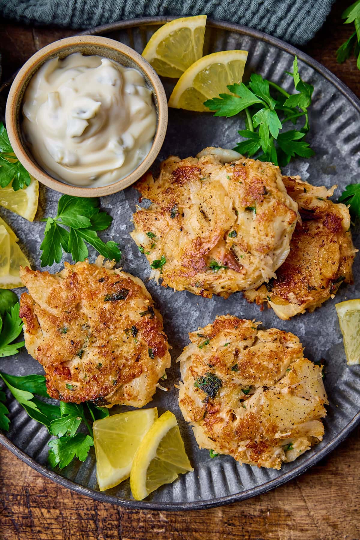 Overhead tall image of a plate of Maryland-style crab cakes on a matte steel plate. There is a small bowl of tartar sauce, some lemon slices and fresh parsley also on the plate. The plate in on a wooden background.