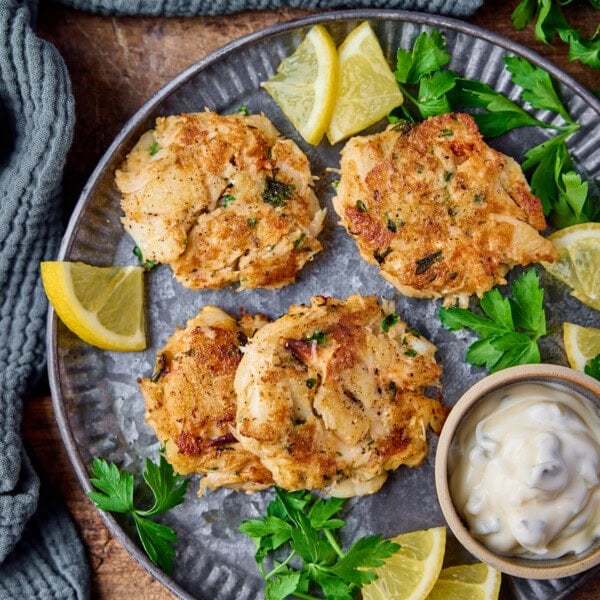 Overhead sqaure image of a plate of Maryland-style crab cakes on a matte steel plate. There is a small bowl of tartar sauce, some lemon slices and fresh parsley also on the plate. The plate in on a wooden background and there is more parsley and a light green napkin near the plate.