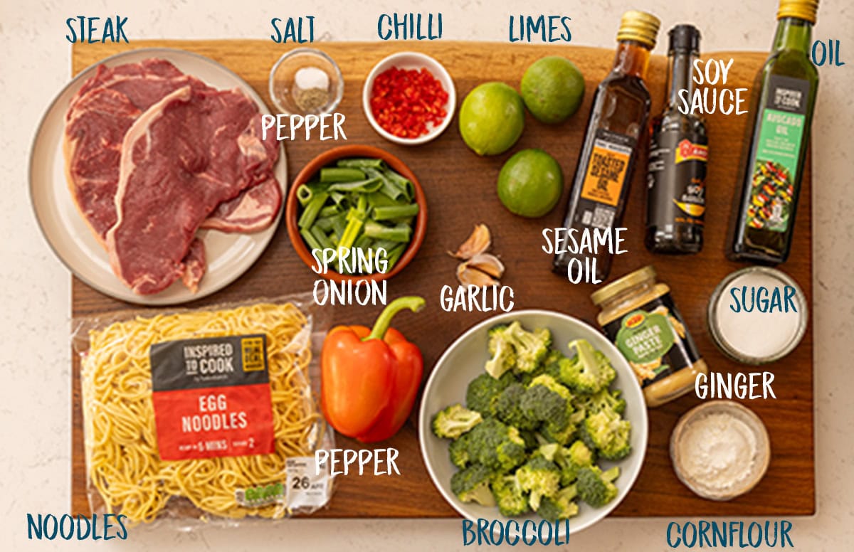 An overhead shot of the ingredients for Crispy Chilli Beef Noodles is set out on a wooden board. They are all labelled in white or dark blue text. They are as follows: Steak, Noodles, Salt, Pepper, Spring Onions, Pepper, Chilli, Garlic, Broccoli, Limes, Sesame Oil, Soy Sauce, Oil, Ginger, Sugar, and Cornflour.