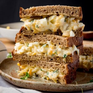 Square side-on image of 3 egg mayonnaise sandwich halves, on brown bread, piled up on a brown plate. The plate is on a light coloured table next to a light striped napkin. The background of the image is black.