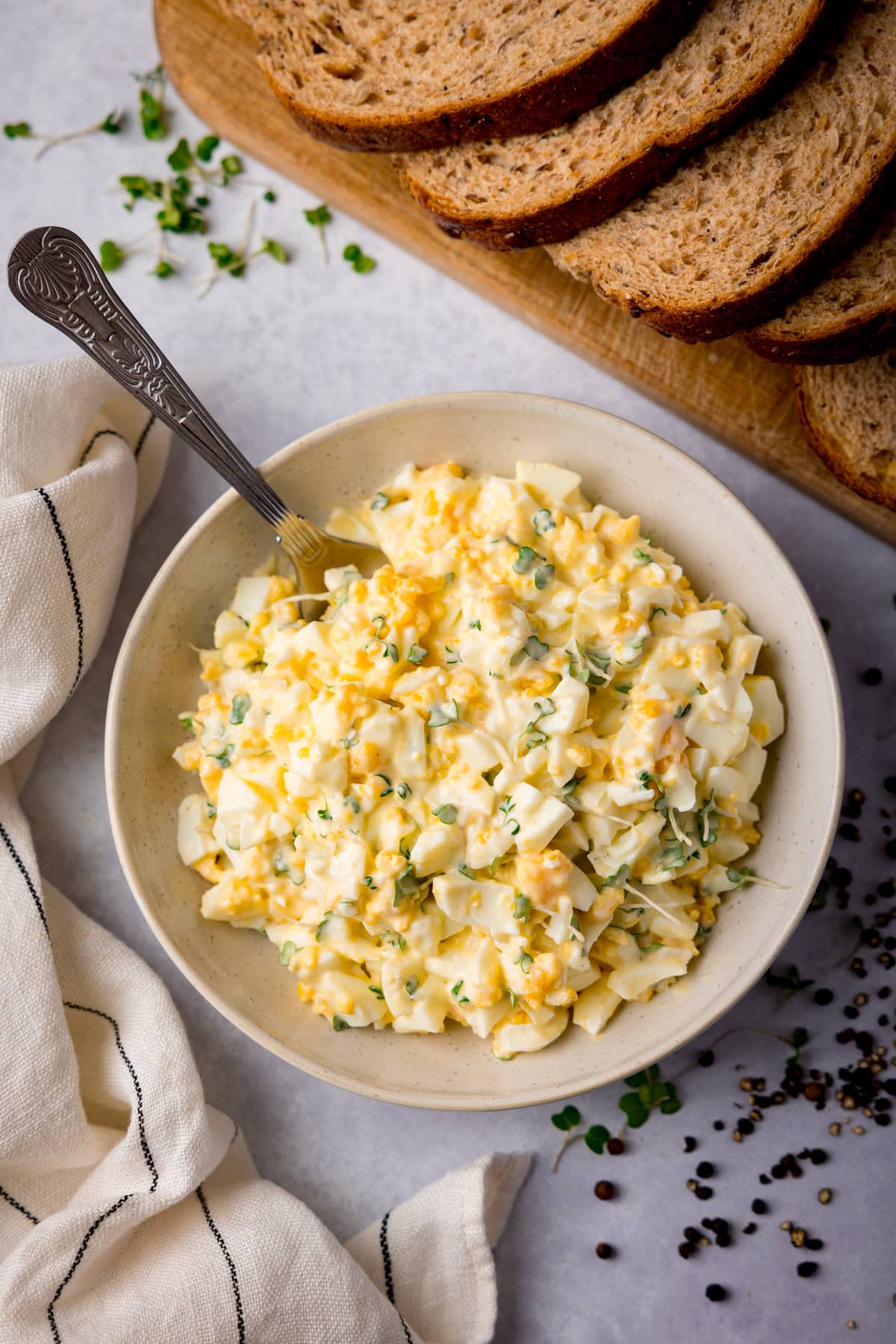 Tall overhead image of egg mayonnaise in a white bowl with a silver spoon sticking out. The bowl is on a light surface and there is a light stripy napkin next to it. There is cress and peppercorns scattered around the bowl and a board with brown bread slices at the top of the image.