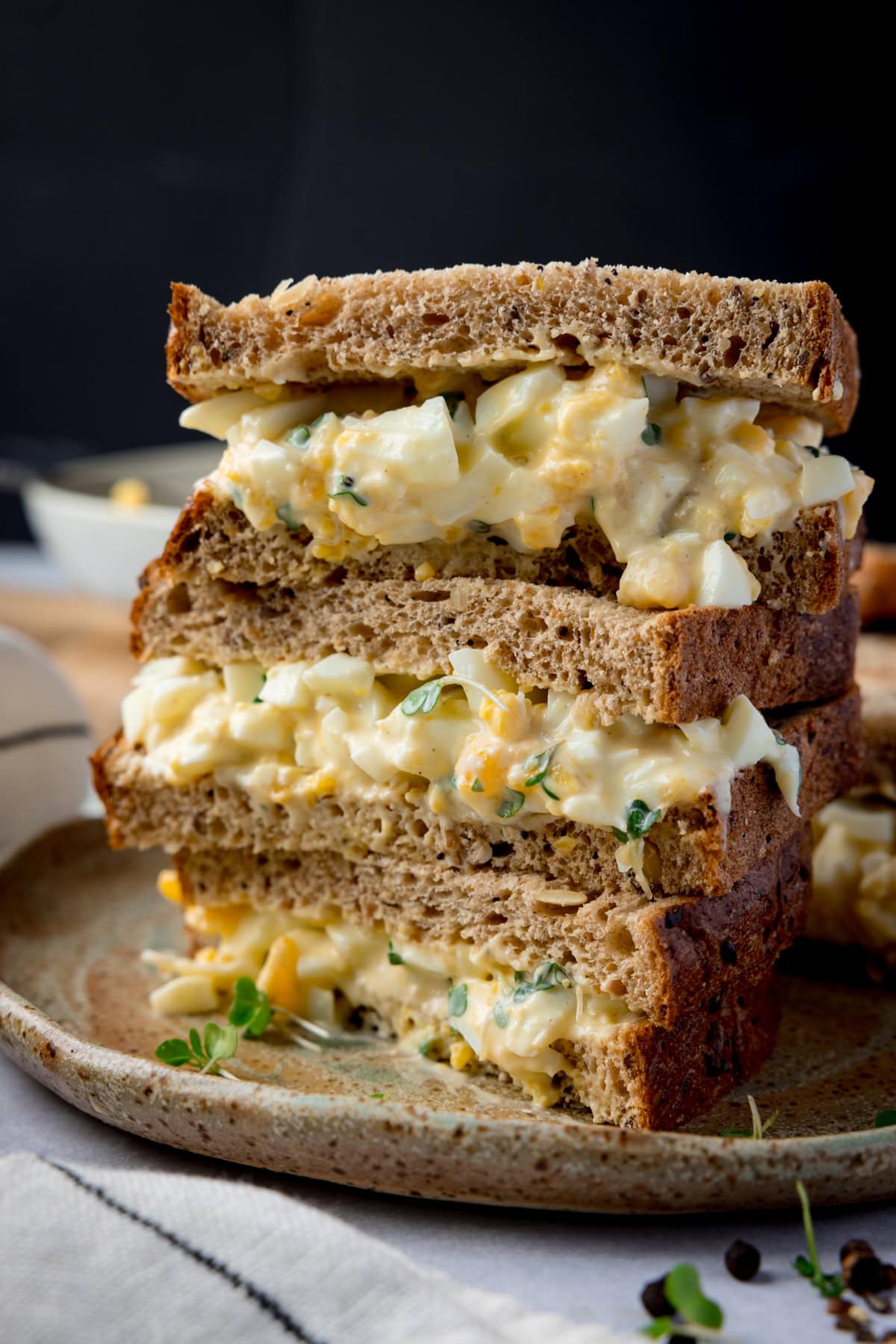 Tall side-on image of 3 egg mayonnaise sandwich halves, on brown bread, piled up on a brown plate. The plate is on a light coloured table next to a light striped napkin. The background of the image is black.