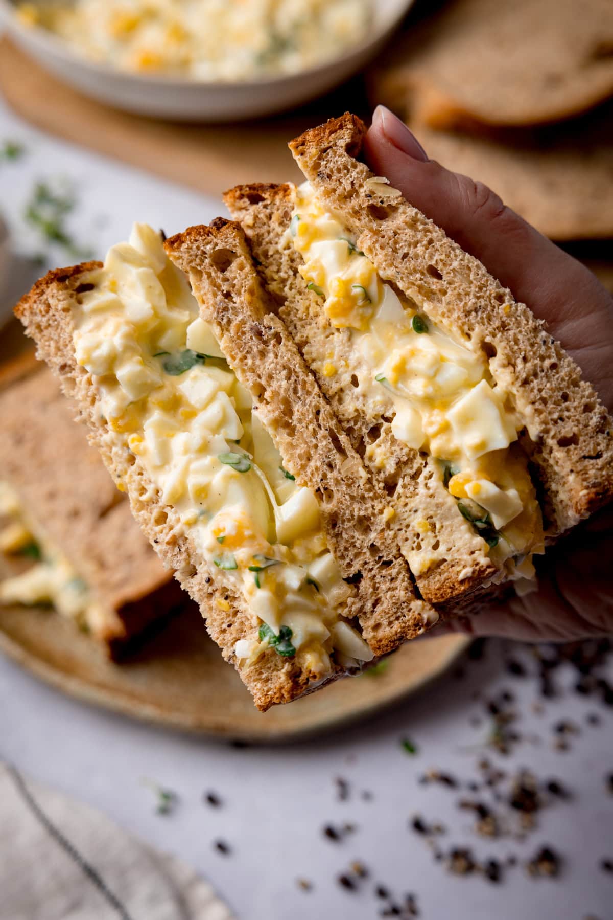 Overhead image of an egg mayonnaise sandwich on brown bread being held to show off the filling. The table underneath has a light grey colour and has ingredients on it, including a bowl of egg mayonnaise, brown bread slices and a further egg mayonnaise sandwich on a brown plate.