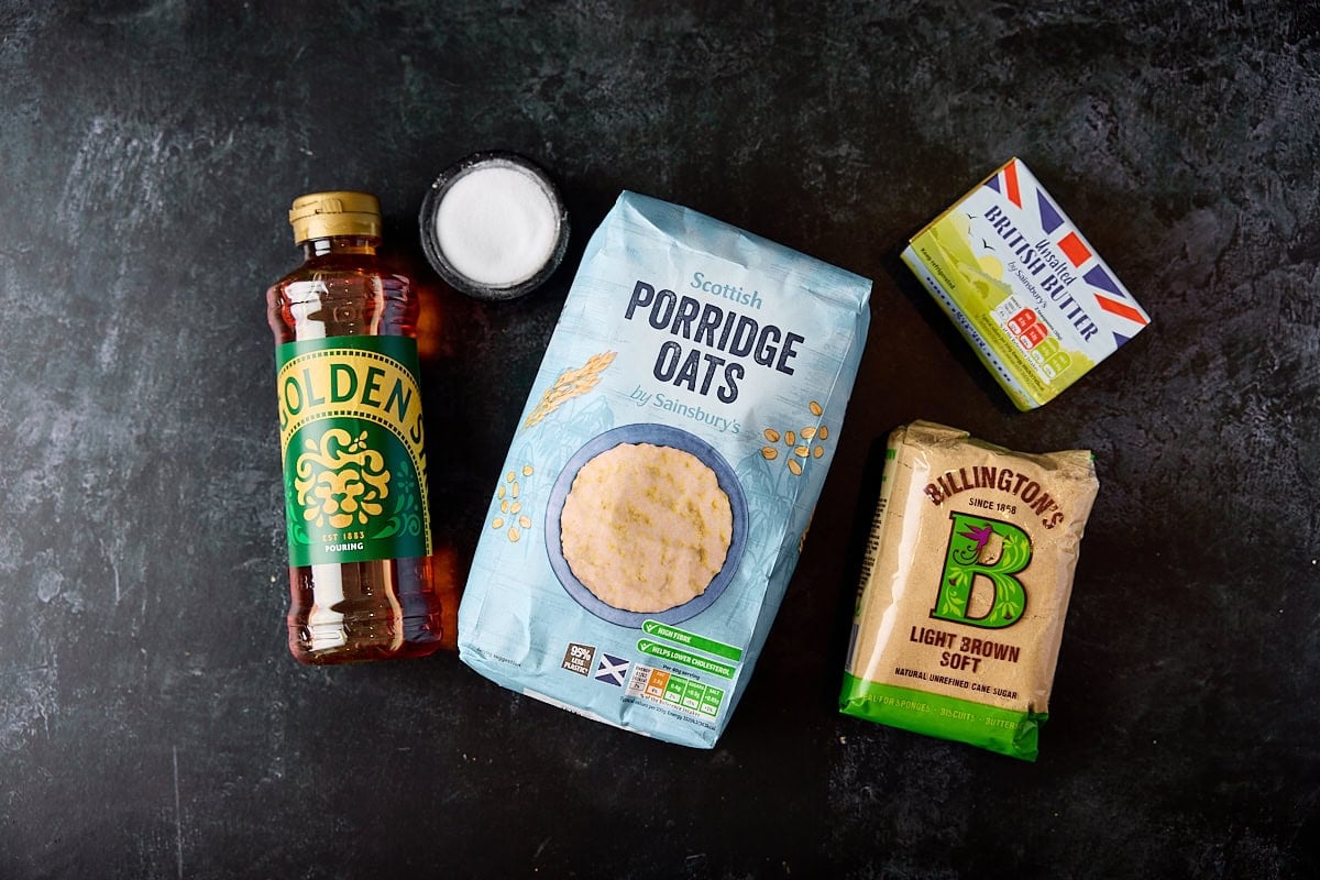 A wide, overhead shot of the ingredients for flapjacks. They are laid out on a black surface. They are as follows: butter, light brown sugar, golden syrup, porridge oats, and salt.