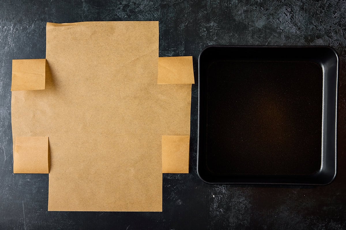 A wide overhead prep shot for the Easy Flapjacks Recipe. The image shows a square black baking tray on the right of the image, and on the left of the image is a sheet of brown parchment paper cut in a specific way to fit the baking tray. This is all set on a black background.