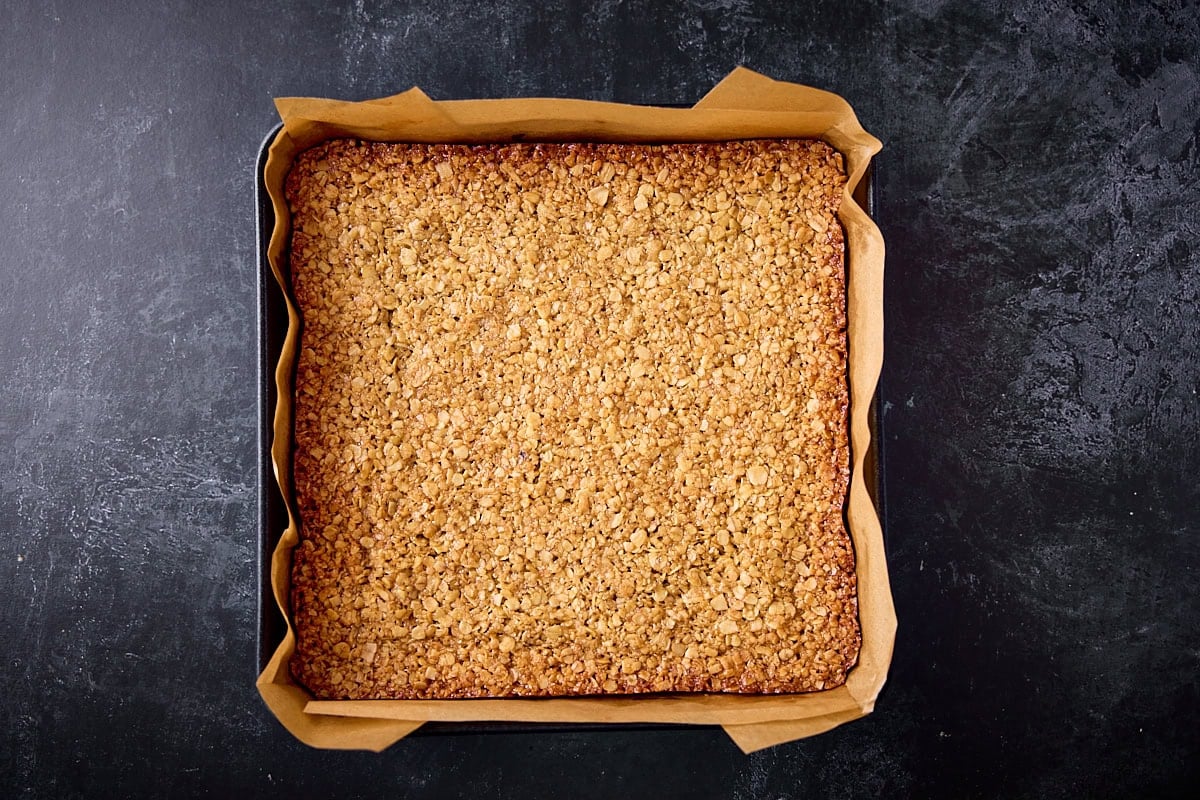 A wide overhead prep shot of flapjacks. The image shows the cooked flapjacks in a black baking tray lined with brown greaseproof paper. This is all set on a black background.