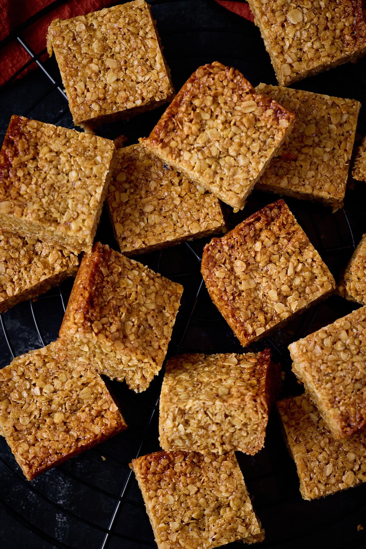 A tall, overhead shot of many flapjack squares scattered around on top of a black wire cooling rack. In the top left of the image, you can slightly see a red/orange napkin tucked beneath the cooling rack. This is all set on a black background. 