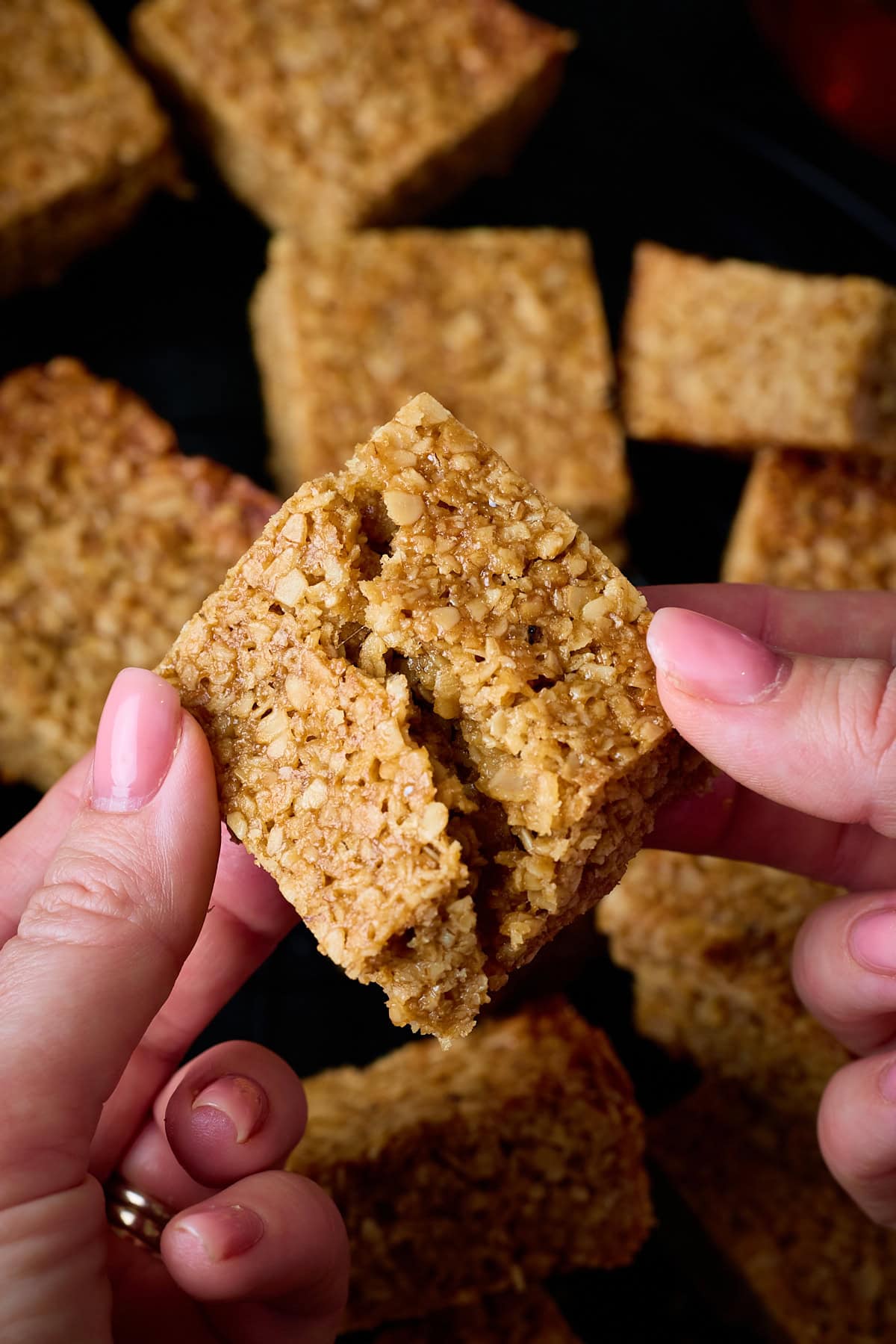 A tall, overhead shot of many flapjack squares scattered around on top of a black wire cooling rack. In the foreground of the image, there are two hands holding one flapjack square and breaking it in half. This is all set on a black background. 