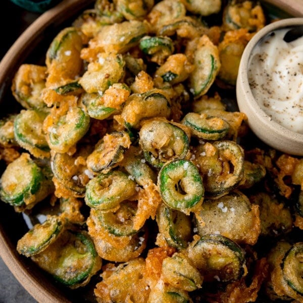 A square, close-up image of a light brown bowl of fried Jalapenos. In the top right of the bowl, there is a light grey dish nestled in the fried Jalapenos, which has garlic mayo in it. In the top left of the background, you can slightly see a dark green napkin. This is all set on a grey surface.