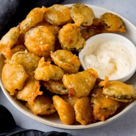 A square image of many fried pickle bites in a white bowl. Nestled in the right of the dish, there is a smaller, white dish filled with garlic mayo. In the left of the background, you can see there is a slate grey napkin tucked underneath the main bowl. This is all on a light grey background.