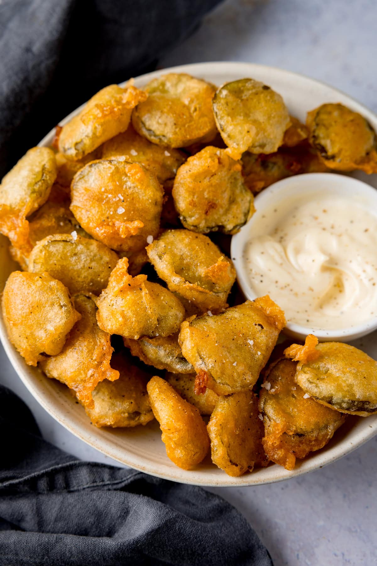 A tall image of many fried pickle bites in a white bowl. Nestled in the right of the dish, there is a smaller, white dish filled with garlic mayo. In the left of the background, you can see there is a slate grey napkin tucked underneath the main bowl. This is all on a light grey background.