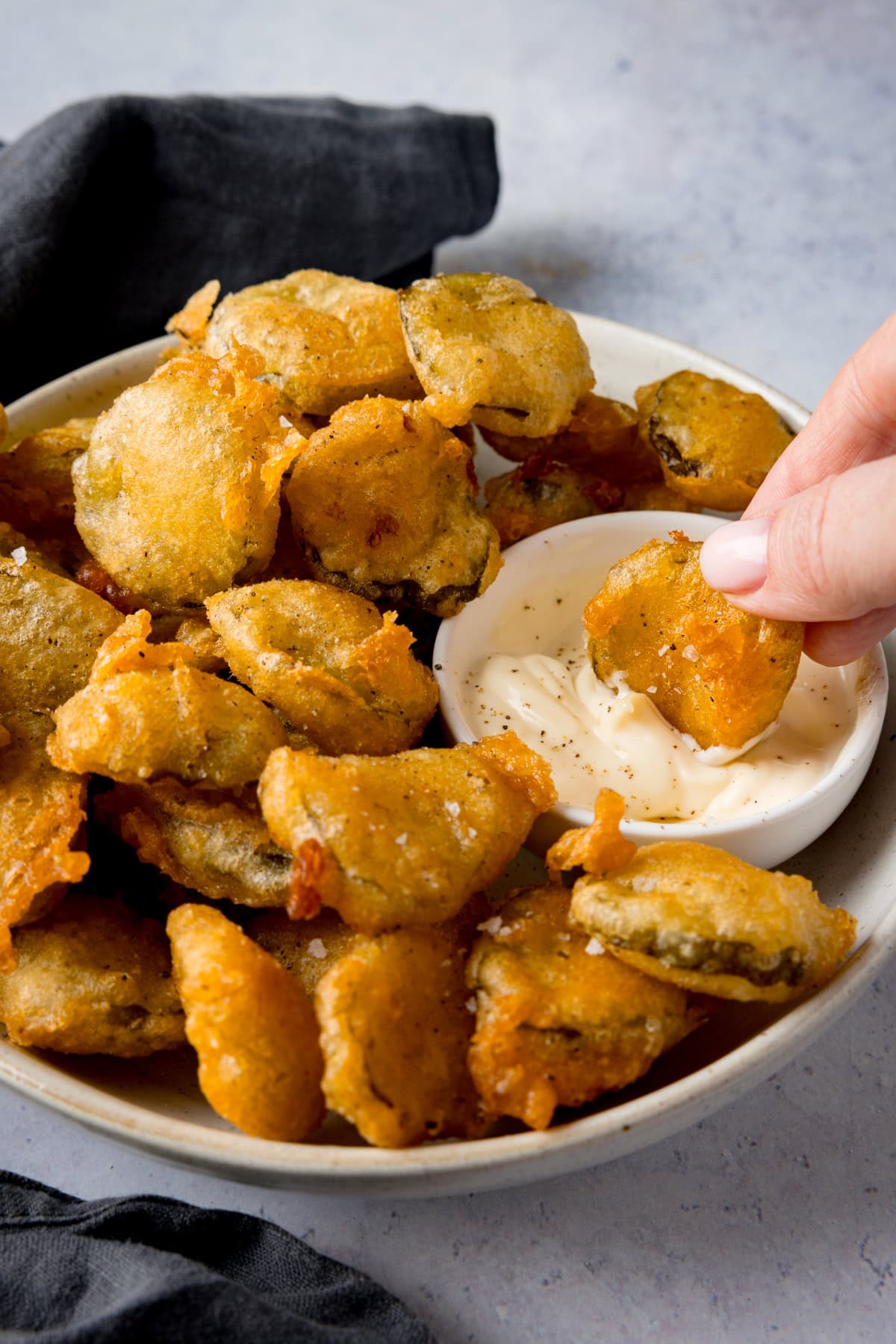 A tall image of many fried pickle bites in a white bowl. Nestled in the right of the dish, there is a smaller, white dish filled with garlic mayo. In the right of the image, there is a hand dipping one of the fried pickles into the dish of garlic mayo. In the left of the background, you can see there is a slate grey napkin tucked underneath the main bowl. This is all on a light grey background.