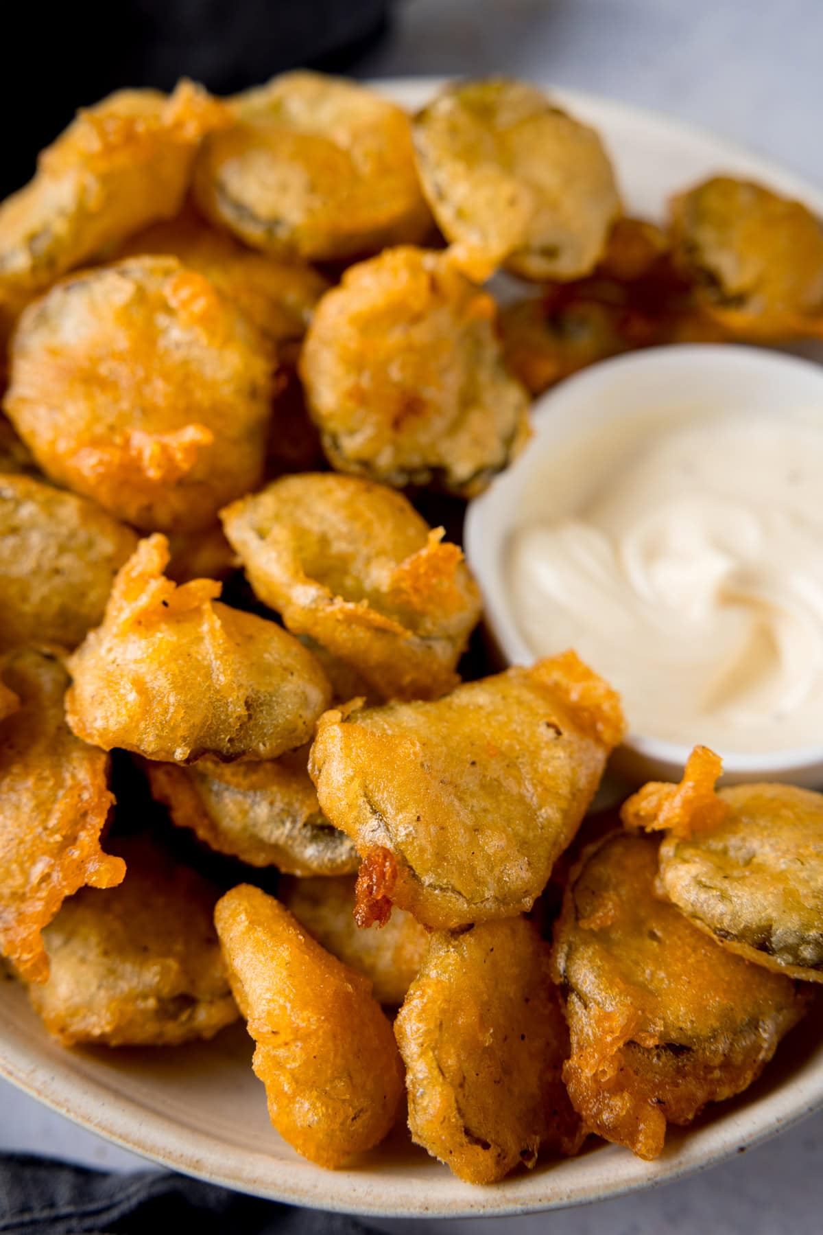 A tall, close-up image of many fried pickle bites in a white bowl. Nestled in the right of the dish, there is a smaller, white dish filled with garlic mayo. In the left of the background, you can see there is a slate grey napkin tucked underneath the main bowl. This is all on a light grey background.