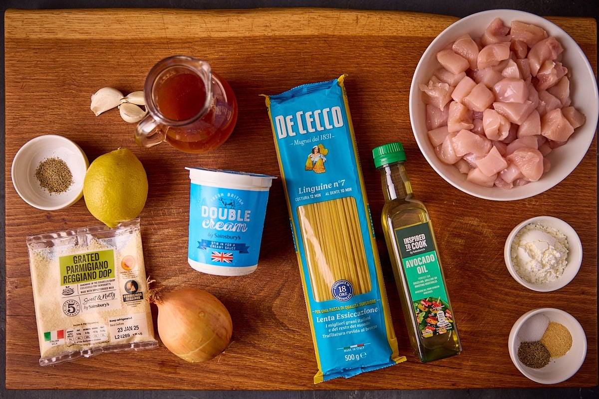 Overhead photo of individual ingredients for making garlic parmesan chicken pasta on a wooden board. The ingredients include, chopped uncooked chicken in a bowl, cornflour, seasonings, oil, pasta, chicken stock, cream, garlic, parmesan cheese, onion and a lemon.