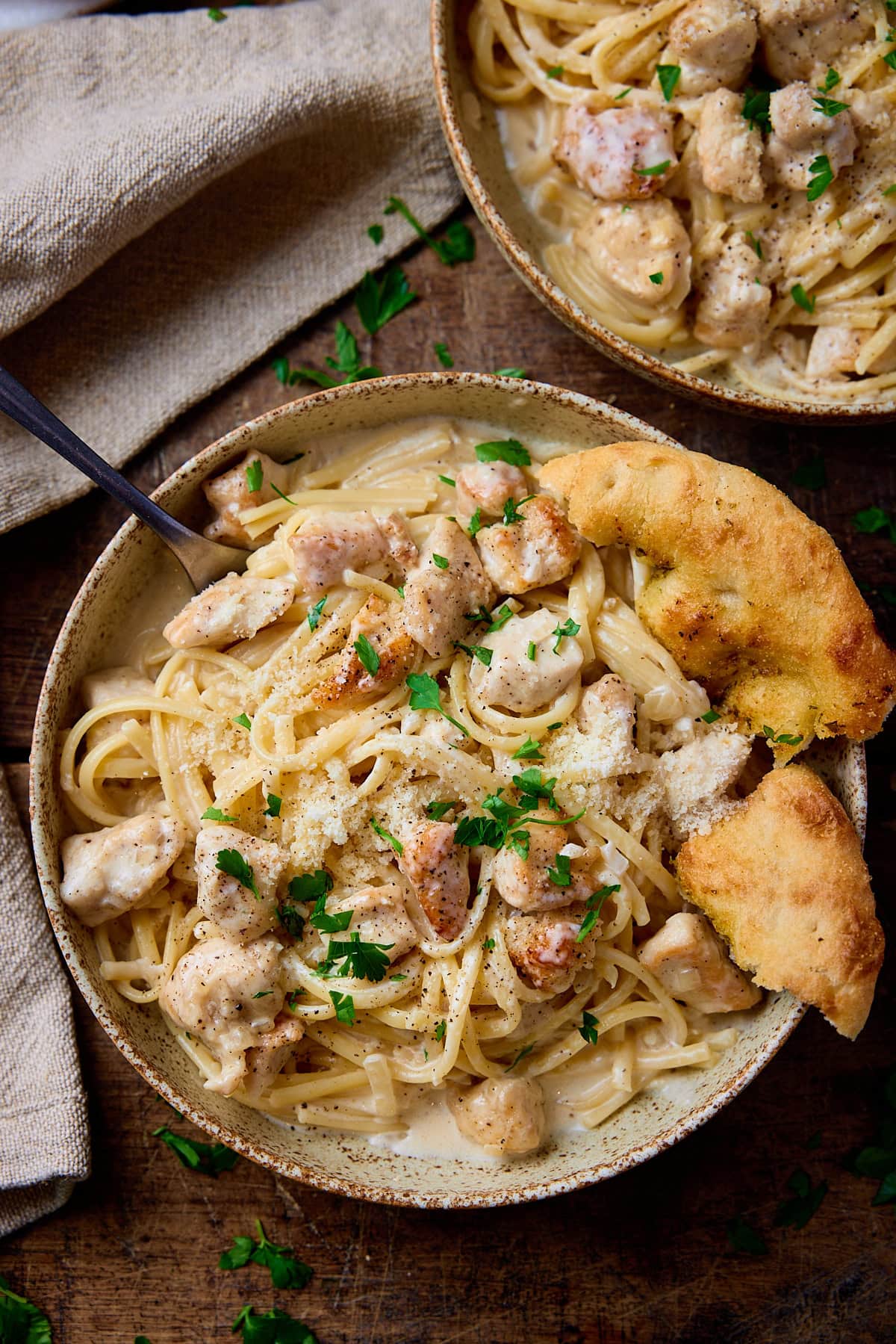 Overhead picture of a stoneware bowl on a wooden board, the bowl is full of pasta and chicken in a garlic parmesan sauce. There is a chunk of garlic bread on the side of the bowl and a fork sticking out of the pasta.