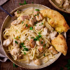 A stoneware bowl on a wooden board, the bowl is full of garlic parmesan chicken pasta. There is a chunk of garlic bread on the side of the bowl and a fork sticking out of the pasta.