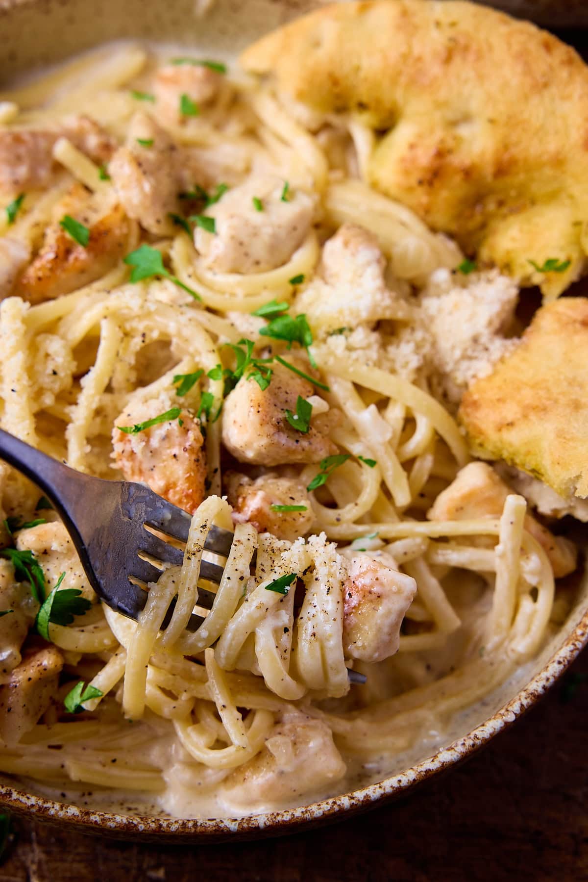 A close up picture of a dark fork full if garlic parmesan chicken pasta. The fork has a swirl of pasta on it and chunk of chicken and is being lifted from the stoneware bowl showing how the lovely creamy sauce sticks to the pasta.