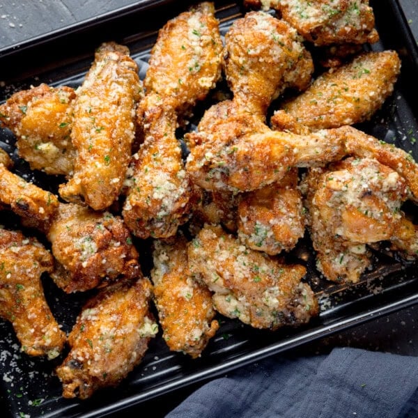 A square, overhead shot of a pile of Garlic Parmesan Chicken Wings on a black rectangular baking tray. In the bottom right corner of the image, you can see a dark grey napkin tucked beneath the tray. This is all set on a dark grey background.