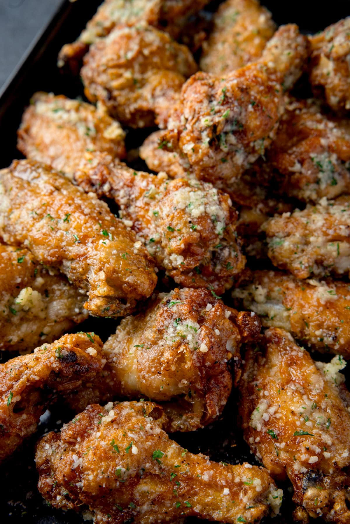 A tall, overhead shot of a pile of Garlic Parmesan Chicken Wings on a black rectangular baking tray. This is all set on a dark grey background.