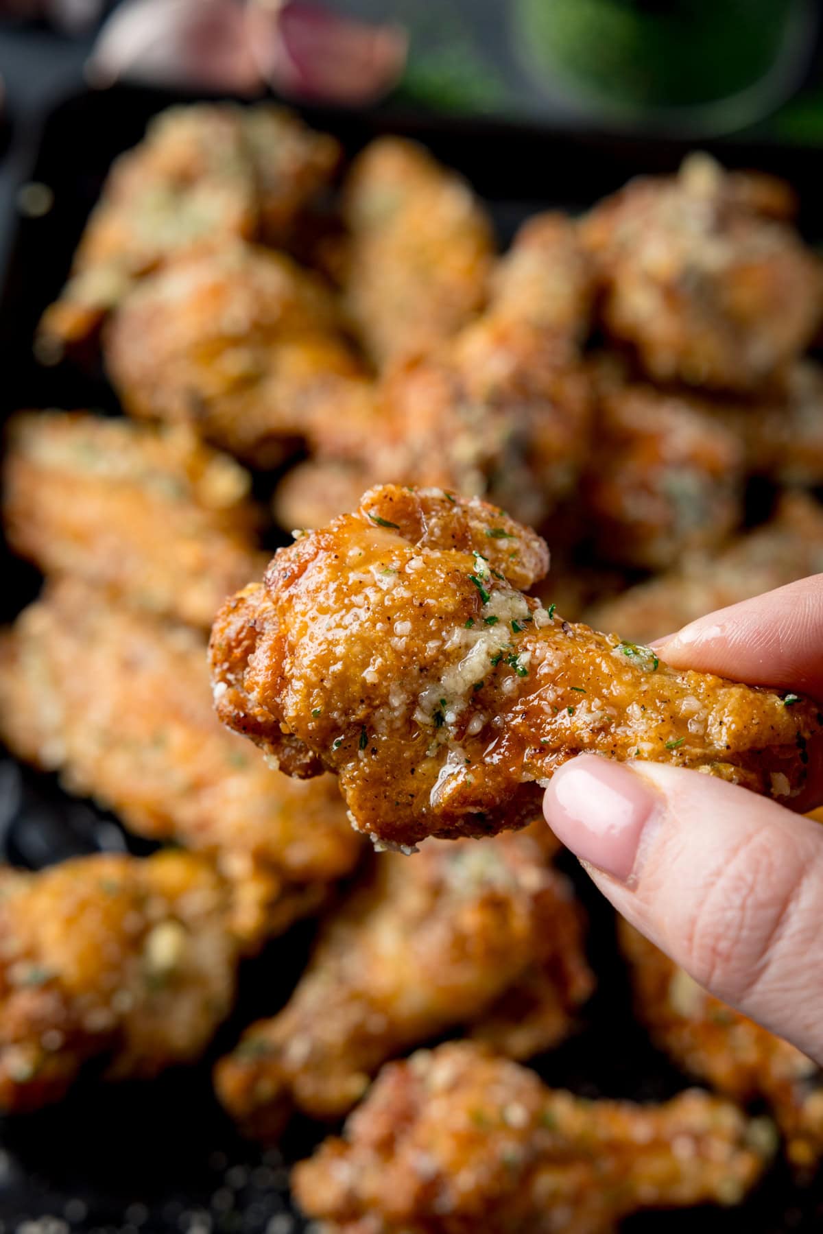 A tall shot of a pile of Garlic Parmesan Chicken Wings on a black rectangular baking tray. Above the baking tray, on the left side. you can see some garlic cloves, and to the right of that, there is a small black dish filled with dried parsley, there is also some dried parsley scattered around it on the surface. In the bottom right corner of the image, you can see a dark grey napkin tucked beneath the tray. In the foreground of the image, there is a hand coming out of the right side of the screen holding a single chicken wing above the tray. This is all set on a dark grey background.