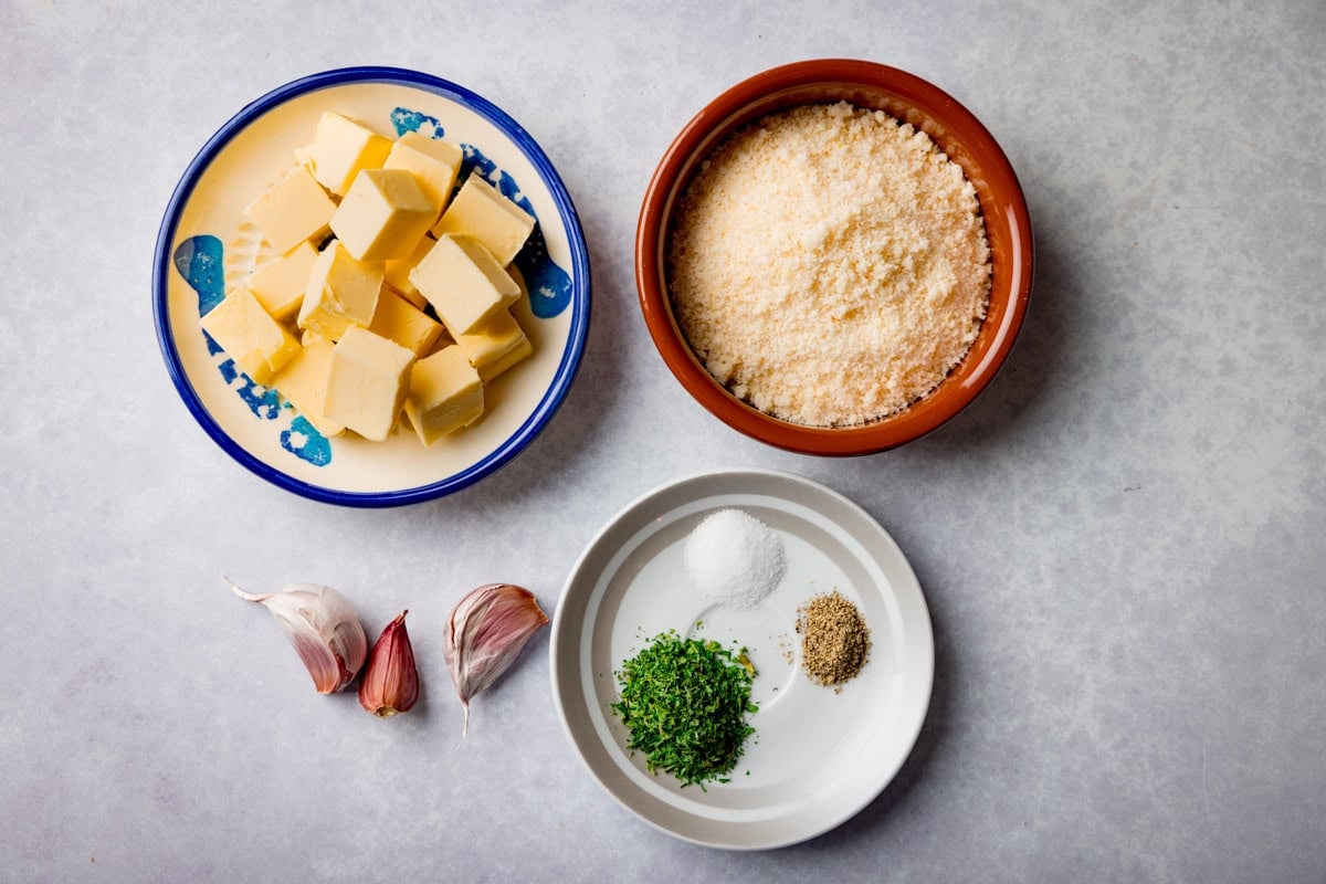 A wide, overhead shot of the ingredients for Garlic Parmesan Chicken Wings. They are laid out on a light grey surface, they are as follows: chicken wings, baking powder, butter, garlic, parmesan, parsley, salt, and pepper. 