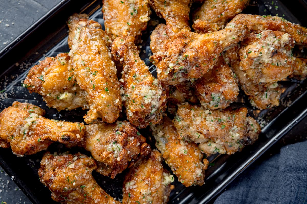 A wide, overhead shot of a pile of Garlic Parmesan Chicken Wings on a black rectangular baking tray. In the bottom right corner of the image, you can see a dark grey napkin tucked beneath the tray. This is all set on a dark grey background.