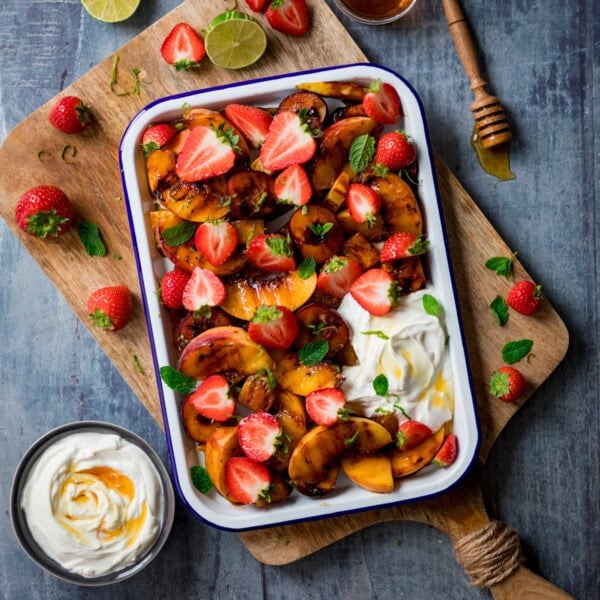 A square, overhead shot of grilled fruit salad in a white tray with a blue lining. There is a dollop of Greek yoghurt on the bottom right side of the tray. The tray is on top of a wooden cutting board, set at an angle. There are strawberries scattered around the wooden board. On the left, below the cutting board, there is a small black bowl, also filled with Greek yoghurt and topped with honey. In the top right corner of the image, you can see a small glass bowl filled with honey, and beside it a honey stirrer, covered in honey. Also, at the top of the image, you can see a few lime wedges, one on the wooden cutting board, and one beside it. This is all set on a light grey background.