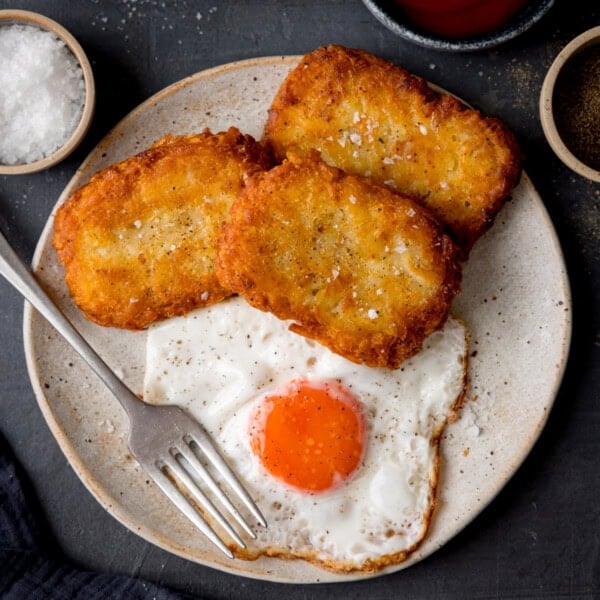 A square, overhead shot of three hash browns on the side of a large white plate, which has a fried egg in the centre, and a silver fork set at an angle on the left-hand side of the plate. In the top left of the image, there is a light brown dish filled with sea salt. In the bottom left of the image, you can slightly see a dark blue napkin. In the top right-hand side of the image, there is a dark blue dish filled with ketchup, and next to that, there is a light brown dish filled with ground pepper. This is all set on a navy blue background.