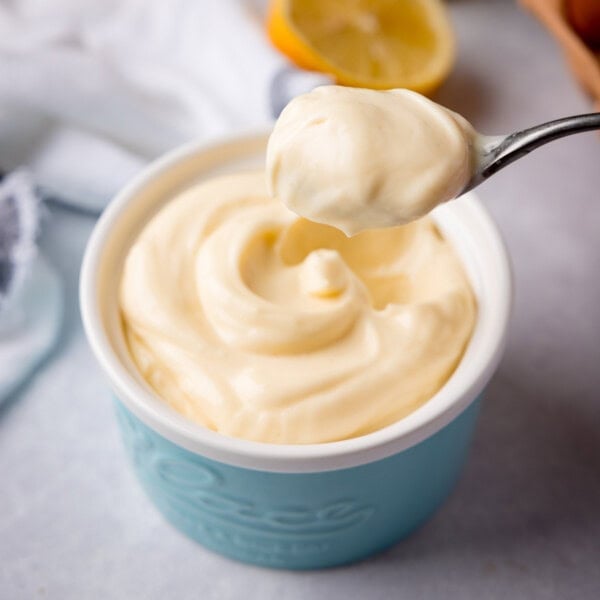 Square image of a small blue ramequin filled with homemade mayonnaise. The ramekin is on a light grey background and there is a white napkin and half a lemon in the background. A spoonful of the mayonnaise is being lifted from the ramequin.