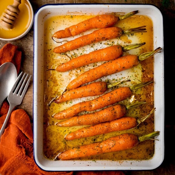 A square, overhead shot of Honey Roasted Carrots in a white tray. To the top left of the tray, there is a small white dish filled with honey, with a wooden honey stirrer in it. Below that, there is an orange napkin with a silver fork and spoon placed on top of it at an angle. This is all set on a wooden surface.