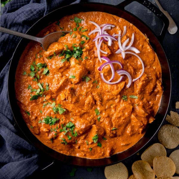 A square, overhead shot of leftover turkey curry in a black skillet. The curry is topped with chopped coriander and red onion, and there is a silver spoon sticking out of the left side of the dish. On the left side of the image, you can see a dark grey napkin tucked beside the pan. In the bottom right corner of the image, you can see some small popadoms scattered around, and in the top right of the image, you can slightly see the handle of a silver fork resting on the surface. This is all set on a black surface.