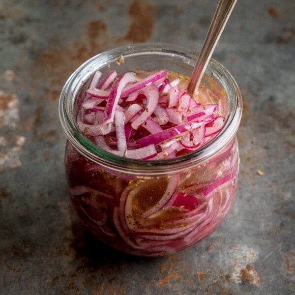 Marinated sliced red onions in a glass jar with a metal spoon sticking out. The jar is on a vintage steel surface.