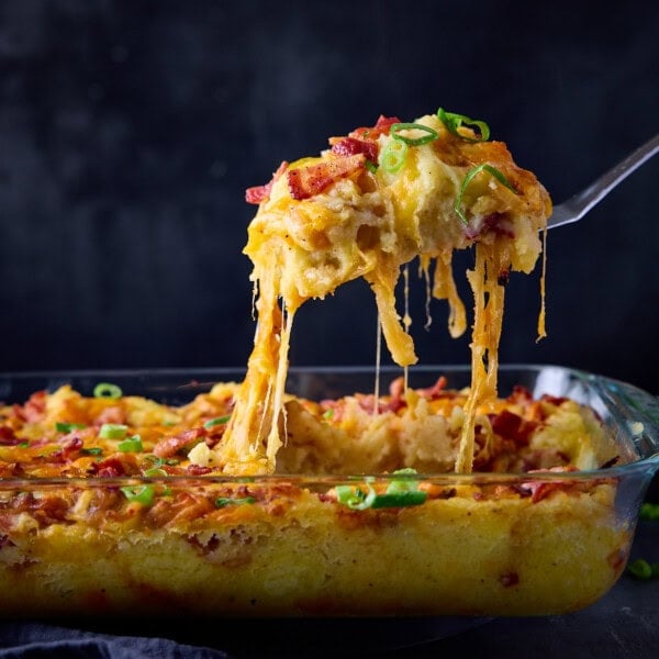 A square image of Mashed Potato Casserole in a rectangular glass dish. There is a silver spoon coming from the left side of the image, which is being held above the dish, scooping out some casserole from the dish. This is all set on a dark blue surface with a dark blue background.