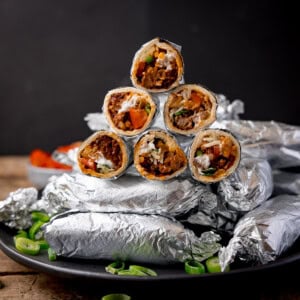 Square image showing mini burritos wrapped in foil, piled up on a plate. Some of the burritos have been sliced in half and piled on top of the whole burritos to show the filling. The plate is on a wooden table against a black background. There is a small bowl of tomatoes blurred out in the background.