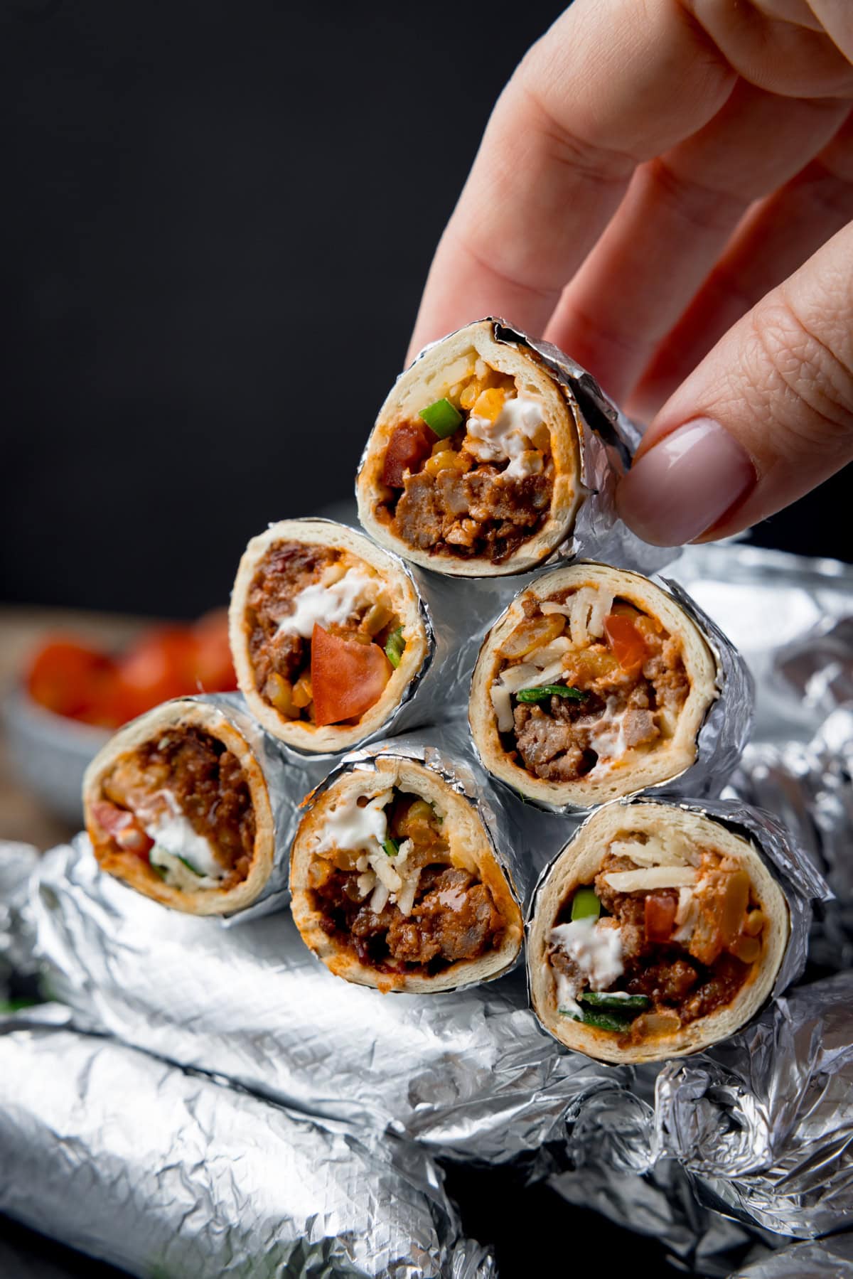 Tall image showing mini burritos wrapped in foil and piled up with a black background. Some of the burritos have been sliced in half and piled on top of the whole burritos to show the filling. A hand is holding picking up one of the open burritos. There is a small bowl of tomatoes blurred out in the background.