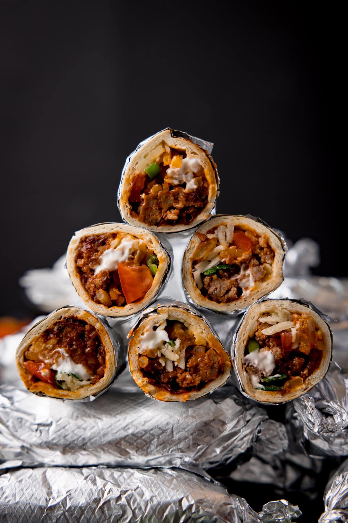 Tall image showing mini burritos wrapped in foil and placed a pile against a black background. Some of the burritos have been sliced in half and piled on top of the whole burritos to show the filling.