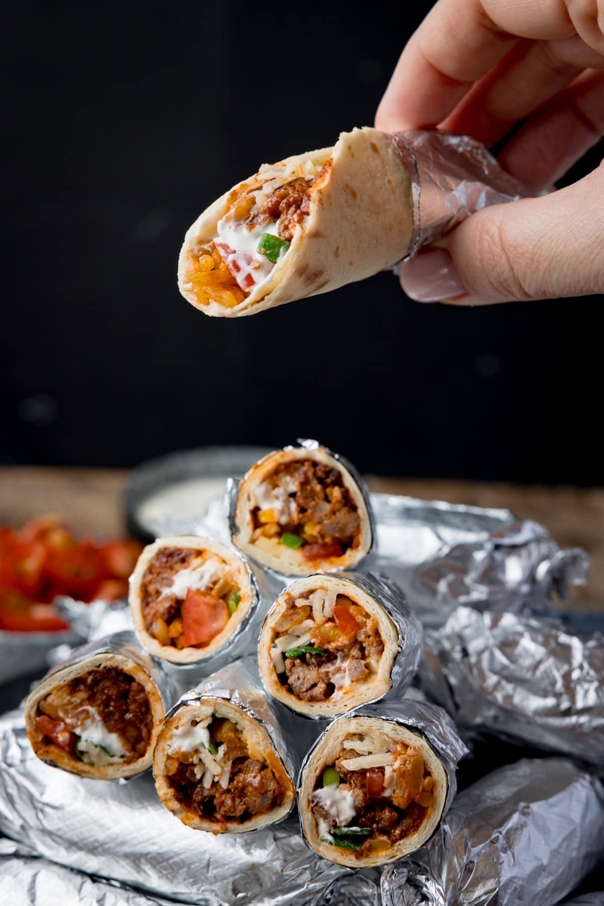 Tall image showing mini burritos wrapped in foil, piled up on a plate against a black background. Some of the burritos have been sliced in half and piled on top of the whole burritos to show the filling. A hand is holding an unwrapped burrito above the pile. There is a small bowl of tomatoes blurred out in the background.