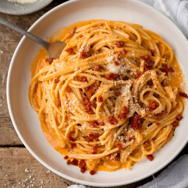 A square, overhead shot of chorizo spaghetti on a large white plate. On the top left side of the plate, there is a silver fork sticking out of the dish. On the right of the background, there is a white napkin tucked beneath the plate. On the top right-hand corner of the image, there is a light grey dish filled with grated parmesan. This is all set on a wooden background.