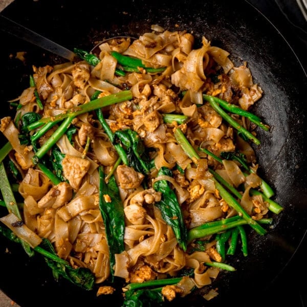 A square, overhead shot of Pad See Ew in a large black wok. In the top left of the wok, you can see the handle of a silver spatula in the dish. This is set on a wooden background.