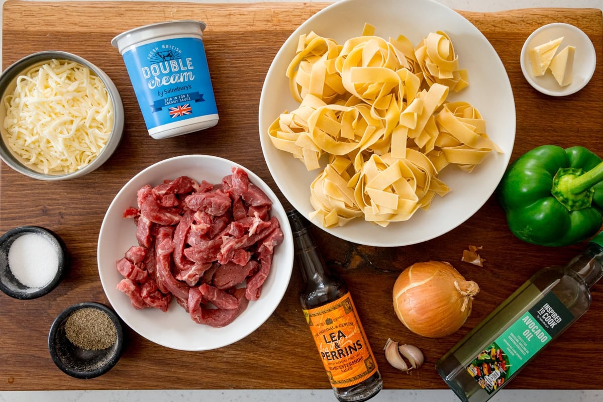 A wide, overhead shot of the ingredients of Philly cheesesteak pasta. They are laid out on a wooden cutting board. The ingredients are as follows: pasta, oil, steak, salt, pepper, butter, onion, green pepper, garlic, double cream, Worcestershire sauce, and grated mozzarella.