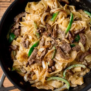 A square, closeup image of Philly Cheesesteak Pasta. The pasta is in the centre of the image, in a black pan with a silver handle. This is all set on a wooden surface.